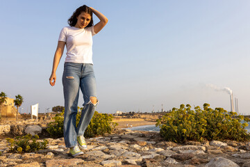 A smiling thoughtful girl in a white T-shirt, blue jeans and light green sneakers walks along the...