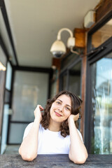 A smiling brunette girl in a white T-shirt, blue jeans sits at a wooden counter in a cafe, resting her chin in her hands. Vertical frame.