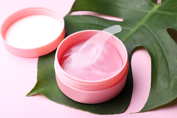 Fototapeta na wymiar Under eye patches in jar with spatula and green leaf on light pink background. Cosmetic product