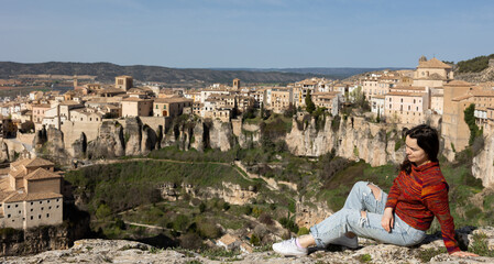 Young adult woman tourist sitting on hill looking at Cuenca old town, Spain