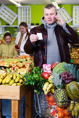Man specifying shopping list by phone while buying fresh fruits and vegetables in shop