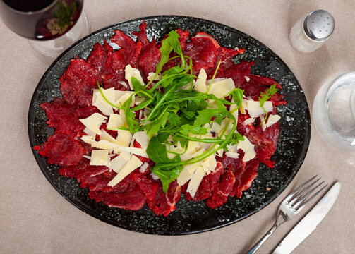 Appetizing fresh meat carpaccio from thinly sliced raw veal with cheese and arugula served on table with glass of red wine. Dish of Mediterranean cuisine