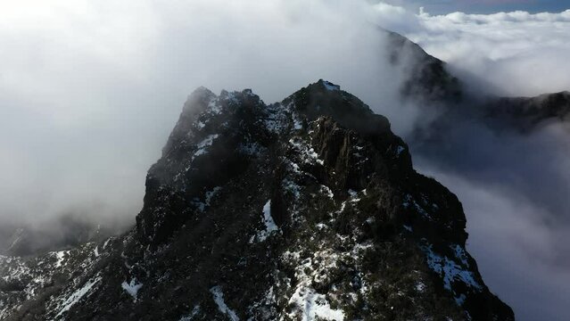 Drone shot of the dark and black peak of the mountain Pico Ruivo in Madeira with thin clouds flying around.