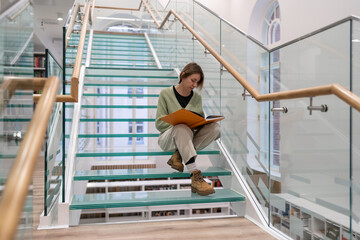 Focused mature female reader sits on modern glass stairs step enjoying interesting book in...