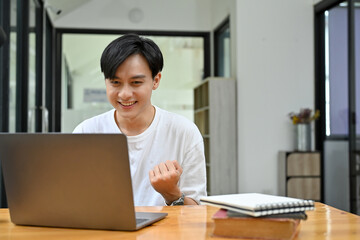 Excited young Asian male looking at his laptop screen, getting an unexpected email