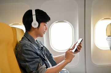 Handsome Asian male traveler at the window seat in economy class is using his mobile phone