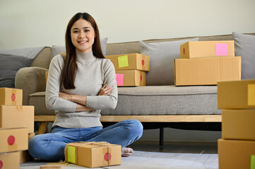 Confident young Asian female small business owner sits in her living room with shipping boxes.