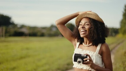 4k Portrait of attractive african-american woman in hat making photos on retro camera in the summer field. Countryside and farmland style.