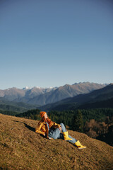 Fototapeta premium Woman full-length sitting resting on a hill and looking at the mountains in a yellow raincoat and jeans happy camping trip in the winter, freedom lifestyle 