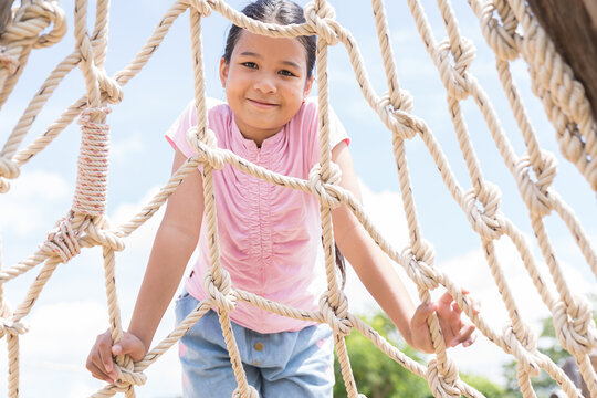 Teenage girl playing on rope wall at the playground. Asian child girl climbing on rope net wall at the adventure rope park - Powered by Adobe