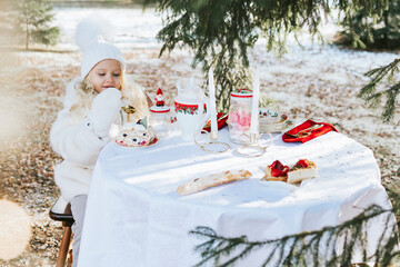 cute little girl in beautiful winter clothes sit at table covered with white tablecloth for...