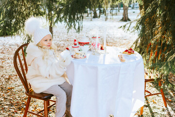 cute little girl in beautiful winter clothes sit at table covered with white tablecloth for breakfast outside in winter, tea, croissants and cakes, a romantic picnic date