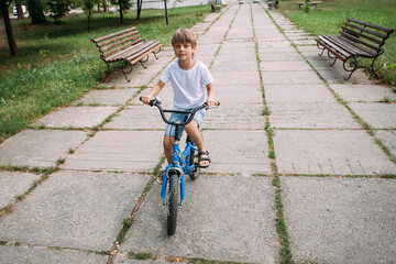 Obraz premium A little cute boy in denim shorts and a white t-shirt rides a children's bike along a path in the park during the day.