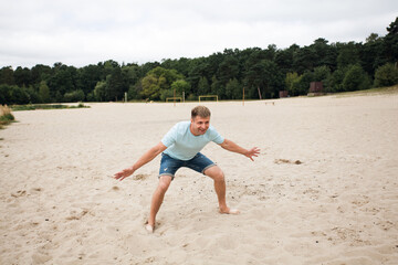 A smiling man in denim shorts and a blue t-shirt stands on the sand, on the beach, like a goalkeeper and is about to catch a soccer ball.