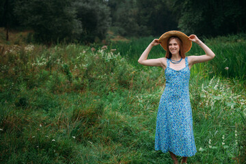 A slender young girl with blond hair, in a blue summer dress and hat, stands on a green lawn in a...