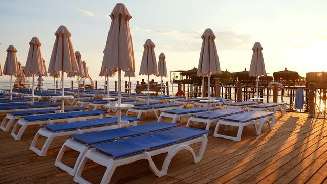 Sun umbrellas and beach chairs on wooden empty beach. Pier and sea in resort. Summer vacation in nature. Tourism and traveling. Rest and relaxation in recreation area to take sun rays and sunbathing.