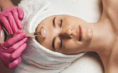 Top view of a caucasian woman getting massage with jade face roller in spa salon. Freckled face....
