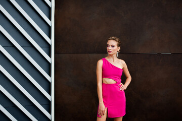 A chic girl with blond hair in a short sexy pink dress, beautiful makeup and a stylish hairstyle is standing on the street against the background of a textured wall of a modern building.
