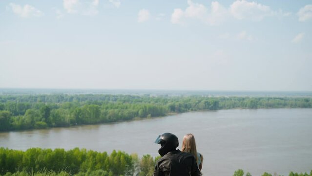 Biker in helmet hugs blonde girlfriend sitting together on motorcycle on green hill top near tranquil lake on spring day backside view slow motion
