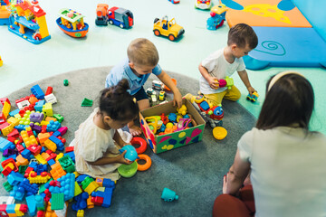 Toddlers and their nursery teacher playing with plastic building blocks and colorful car toys in a...
