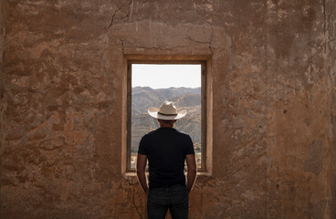 Fototapeta na wymiar Rear view of adult man in cowboy hat standing behind window looking at view