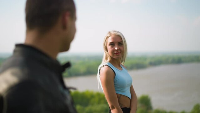 Pretty woman turns head and smiles to beloved man motorcyclist at romantic date on hilly bank of large river on sunny spring day slow motion