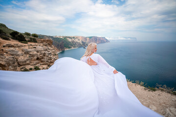 Blonde with long hair on a sunny seashore in a white flowing dress, rear view, silk fabric waving in the wind. Against the backdrop of the blue sky and mountains on the seashore.