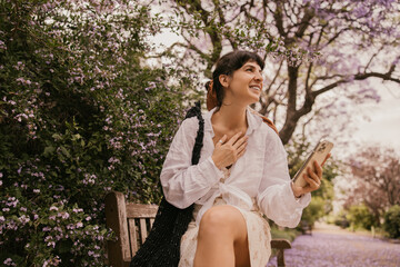 Naklejka premium Pretty caucasian young lady holding smartphone, looking away sitting on park bench. Brunette wears sundress, shirt and bag. Concept lifestyle, technology