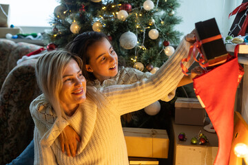 Merry Christmas and Happy Holidays. Cheerful mom and her cute daughter girl exchanging gifts. Parent and little child having fun near Christmas tree indoors. Loving family with presents in room.