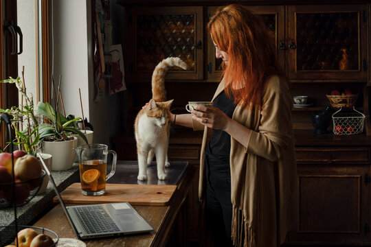 Woman drinking tea and stroking cat on the kitchen. Cozy morning routine at home. Cute ginger cat with owner on the table. Vintage home, stylish old-fessioned house life. Pet friend