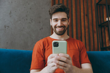 Young fun man in red t-shirt hold in hand use mobile cell phone sit on blue sofa couch stay at home...