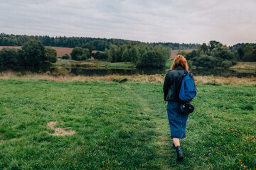 Naklejka na ściany i meble Young woman walking outdoors in green autumn field in evening. Woman with a backpack hiking outdoors