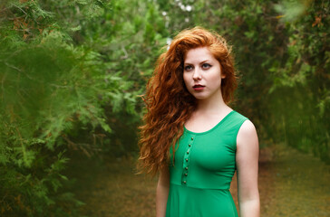 Portrait red beautiful young woman, against green summer park. In green dress with long wavy hair and red lipstick posing