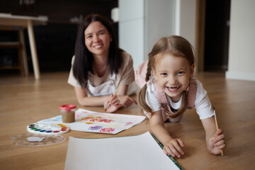 Obraz na płótnie Canvas Smiling young mother and little daughter lie on a warm wooden floor in the living room, drawing together, happy mother rejoices at her daughter's drawings. paint on canvas