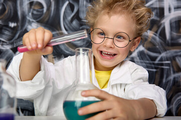 In lab coat, a scientist boy with round glasses conducts an experiment in a laboratory or a chemistry room. Happy child plays with chemical experiments, mixes colors in a test tube. 