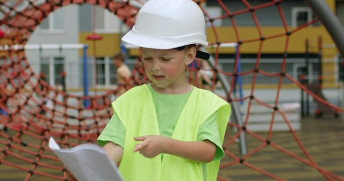 Preschooler boy enjoys playing builder on playground looking at papers. Excited child wearing special uniform and white helmet stands against yard