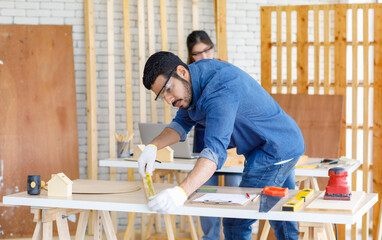 Indian professional male engineer architect foreman labor worker wears safety goggles using measuring tape measure wood plank on working table while female colleague typing laptop computer behind