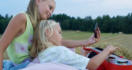 charming girl playing the ukulele on mown rye in field. Landscape of straw bales against setting sun on background. Happy sisters watch video on smartphone outdoors. Childhood. Country life