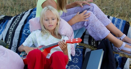 Obraz premium charming girl playing the ukulele on mown rye in field. Landscape of straw bales against setting sun on background. Happy sisters watch video on smartphone outdoors. Childhood. Country life