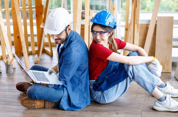 Asian professional female and Indian bearded male engineer architect foreman labor worker wear safety goggles and gloves sitting on floor smiling working with laptop computer discussing together