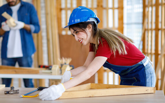 Asian professional female engineer architect foreman labor worker wears safety hard helmet jeans apron and gloves standing using squard angle degree ruler and pencil measuring wood plank on workbench