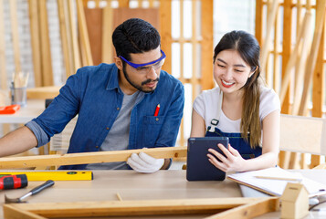 Asian professional cheerful female engineer architect foreman labor worker sitting smiling holding tablet computer discussing with Indian bearded male carpenter in workshop housing construction site