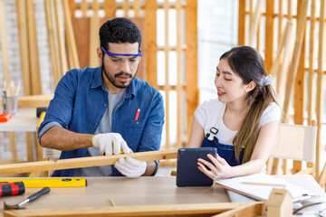 Asian professional cheerful female engineer architect foreman labor worker sitting smiling holding tablet computer discussing with Indian bearded male carpenter in workshop housing construction site