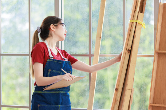 Asian professional female engineer architect foreman labor worker wears safety goggles glasses and jeans apron holding paper clipboard and pencil using hand checking wooden plank spec and condition