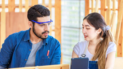 Asian professional cheerful female engineer architect foreman labor worker sitting smiling holding tablet computer discussing with Indian bearded male carpenter in workshop housing construction site