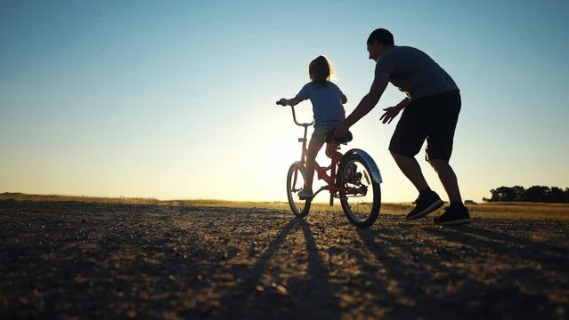 dad teaches daughter to ride a bike. happy family childhood dream concept. father and little daughter learn to ride a bike silhouette in the park. happy family goes in for sports sunlight outdoors