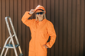 Stylish woman in orange suit, hardhat and sunglasses next to stairs on brown background