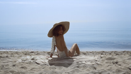 Stylish woman turning back sitting sunny seashore. Curly girl sunbathing beach 