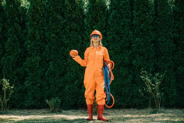 Stylish woman in orange suit, hardhat, pipes and sunglasses with pumpkin on green grass and trees background