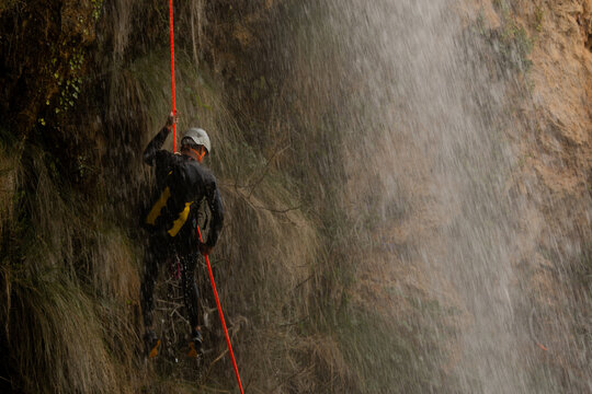 Man wearing helmet and wetsuit rappelling in the middle of a waterfall